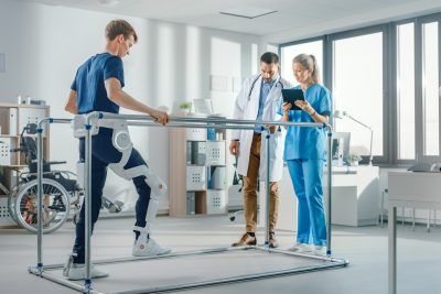 Modern Hospital Physical Therapy: Patient with Injury Walks Wearing Advanced Robotic Exoskeleton. Physiotherapy Rehabilitation Scientists, Engineers use Tablet Computer to Help Disabled Person